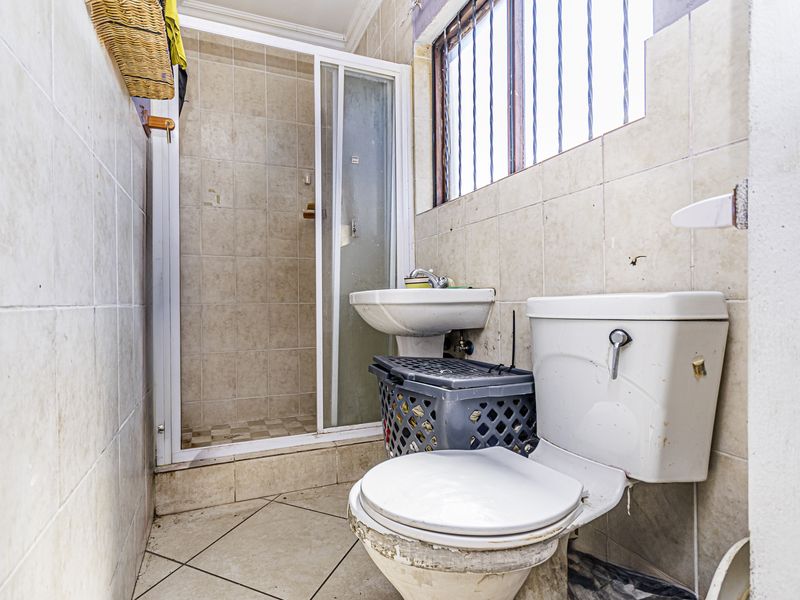 A functional bathroom featuring a shower enclosure, pedestal basin, and toilet, with tiled finishes and a window with security bars.