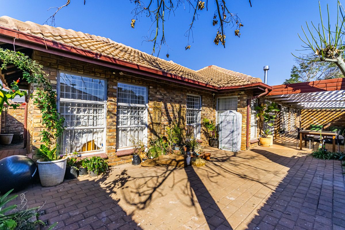 A rear exterior view of a brick house featuring a paved patio area, a pergola, and large windows with security bars.