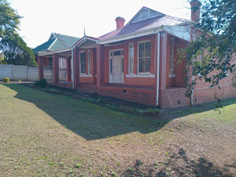 A traditional red brick house featuring a covered veranda, corrugated metal roof, and established garden setting.