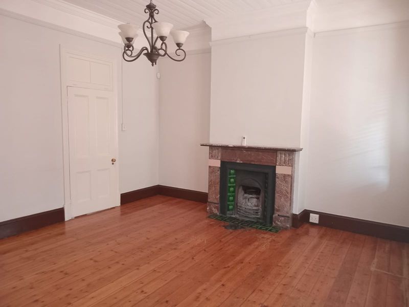 A bright, empty living room featuring original timber flooring, a classic fireplace with mantle, and decorative ceiling cornicing.