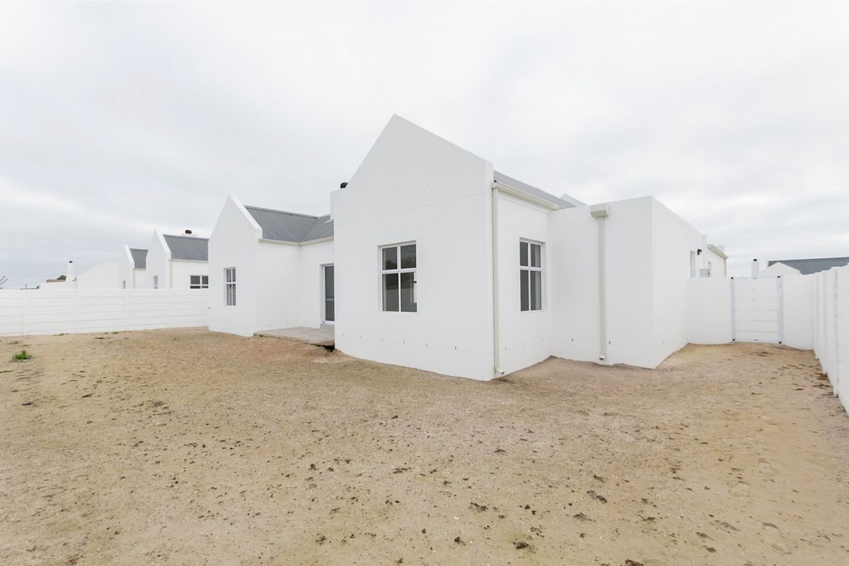 Rear view of a modern white house with a metal roof, featuring a large, unlandscaped sandy yard and perimeter boundary walls.