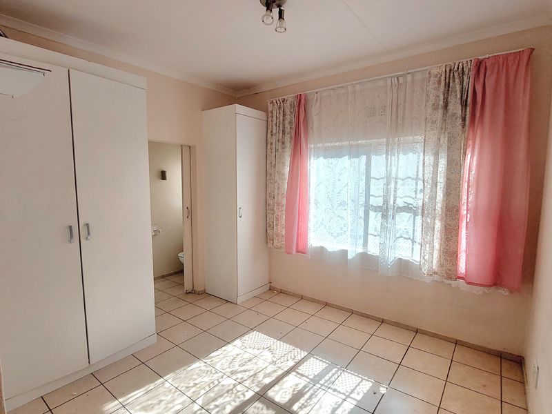 A bright, empty bedroom featuring tiled flooring and built-in cupboards, with an adjoining en-suite bathroom visible through the doorway.