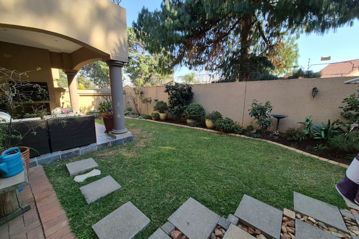 A well-maintained rear garden featuring a covered patio with a built-in braai, stone paving, and a manicured lawn enclosed by a boundary wall.