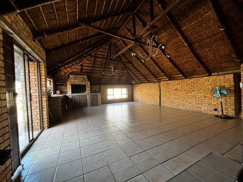 Spacious covered patio featuring a high thatched roof with exposed timber trusses, brick walls, tiled flooring, and a built-in braai area.