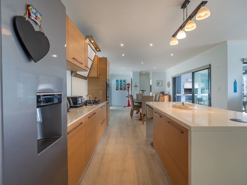 Modern open-plan kitchen featuring timber cabinetry, stone benchtops, and a central island, flowing seamlessly into the dining area and outdoor patio space.