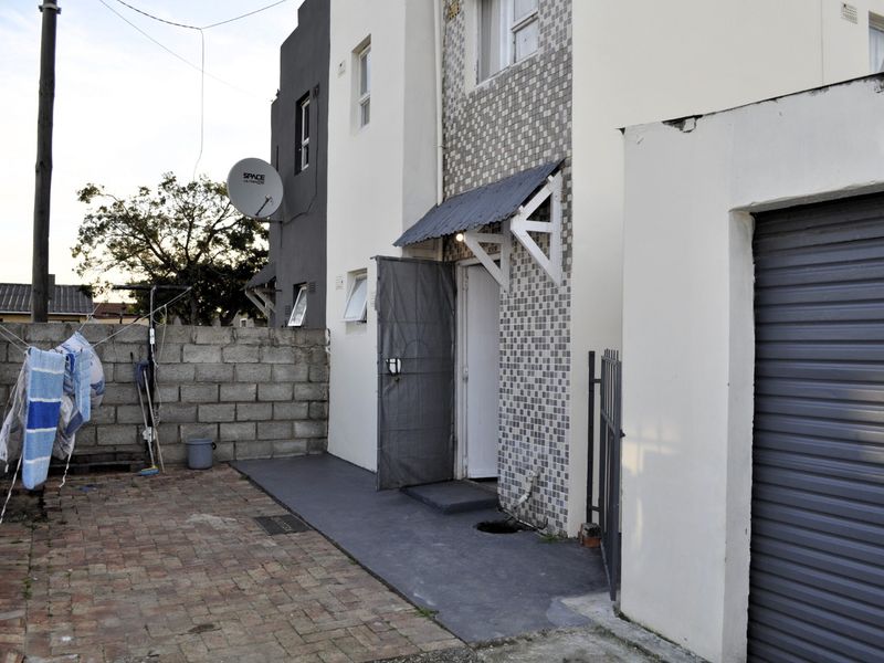 Rear exterior view of the property featuring a paved courtyard, concrete block boundary wall, and a private entrance with a metal awning.