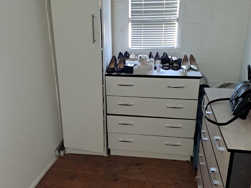 A small bedroom featuring a window with venetian blinds and a ventilation grille, showing signs of wear on the flooring and walls.