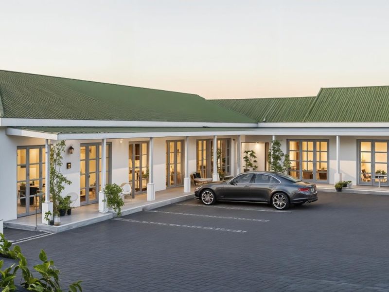 A well-maintained single-story building featuring a green metal roof, a covered veranda with multiple glass doors, and a spacious paved driveway.