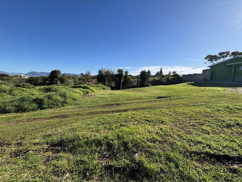 A view of a vacant residential plot featuring a grass lawn and boundary wall, with mountain views in the distance.