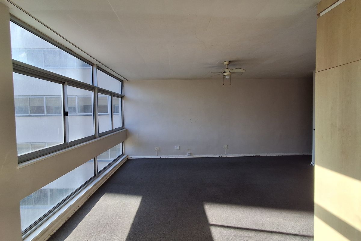An empty, sunlit living area featuring large aluminium-framed windows, a ceiling fan, and built-in cabinetry, offering a blank canvas for renovation.