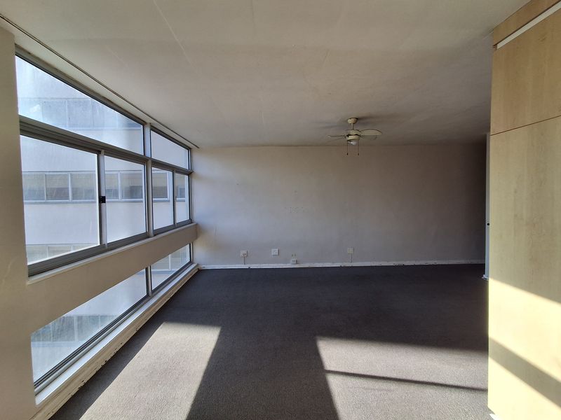 An empty, sunlit living area featuring large aluminium-framed windows, a ceiling fan, and built-in cabinetry, offering a blank canvas for renovation.
