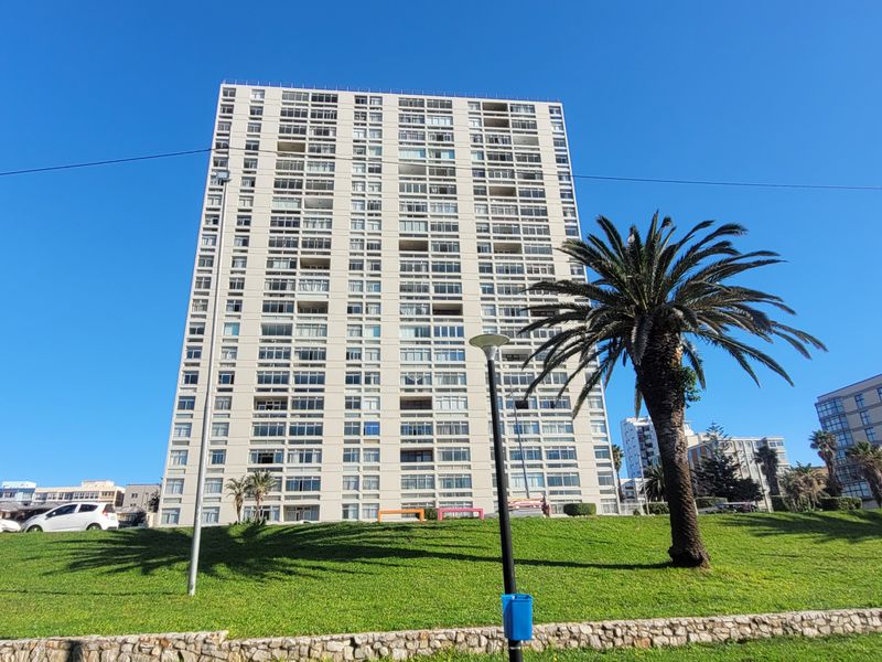 A wide-angle view of a multi-story apartment building facade, featuring balconies and set behind a well-maintained landscaped lawn and stone retaining wall.