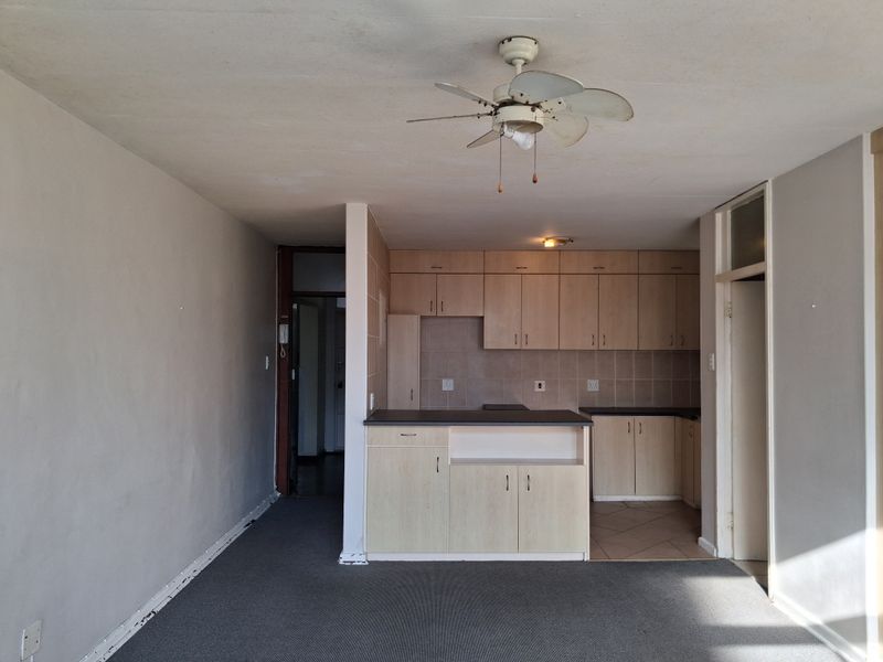 Open plan living area featuring a compact kitchen with built-in cabinetry, tiled backsplash, and a ceiling fan, presenting a dated but functional layout.