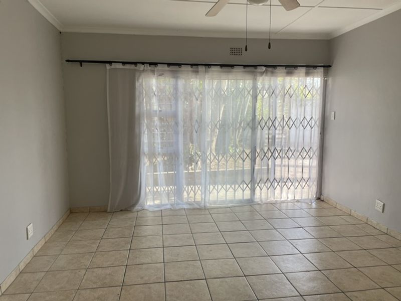 An empty living room featuring neutral tiled flooring, a ceiling fan, and a sliding glass door with an integrated security gate leading outdoors.