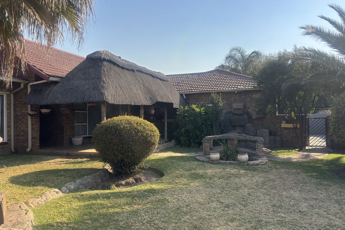 A spacious rear garden featuring a covered thatched patio, brick exterior walls, and a decorative stone water feature set within a landscaped lawn.
