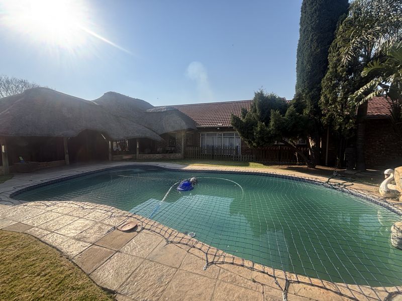 Outdoor pool area featuring a swimming pool with a safety net, paved decking, and a thatched roof structure in the background.