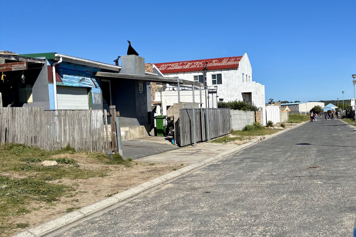 A street-level view of the residential property showing the road frontage, boundary fencing, and adjacent building structures under a clear blue sky.