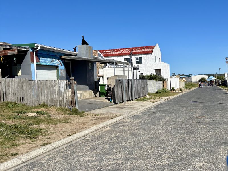 A street-level view of the residential property showing the road frontage, boundary fencing, and adjacent building structures under a clear blue sky.