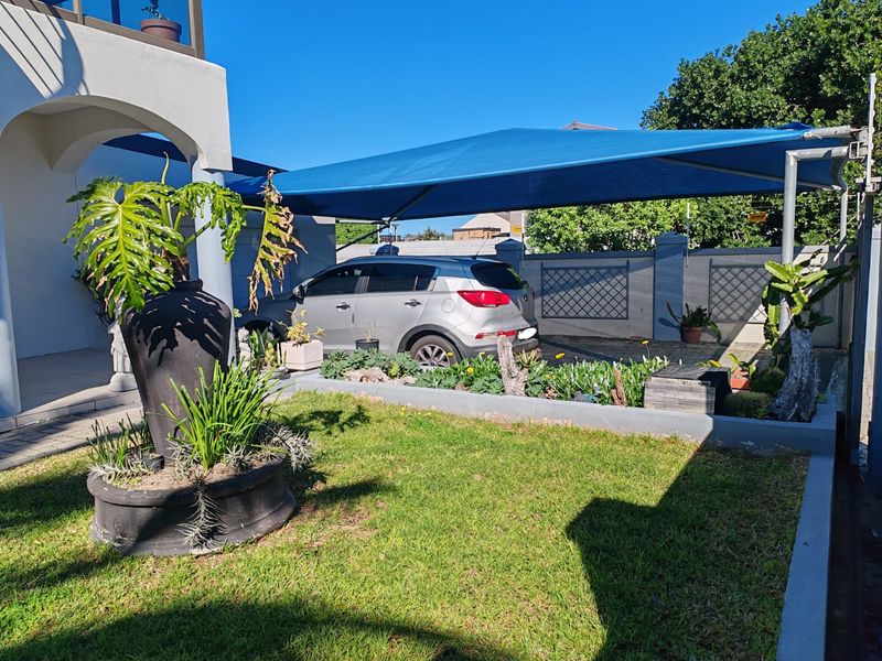 Front exterior view featuring a paved driveway with a blue shade sail, a manicured lawn, and a raised garden bed.