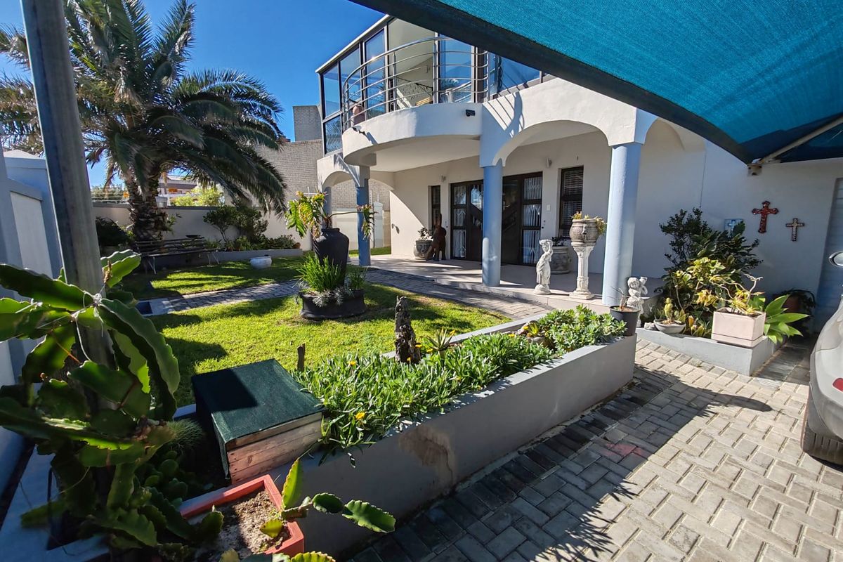 Rear exterior view featuring a landscaped garden, paved driveway, raised planter beds, and a covered balcony with a shade sail structure.