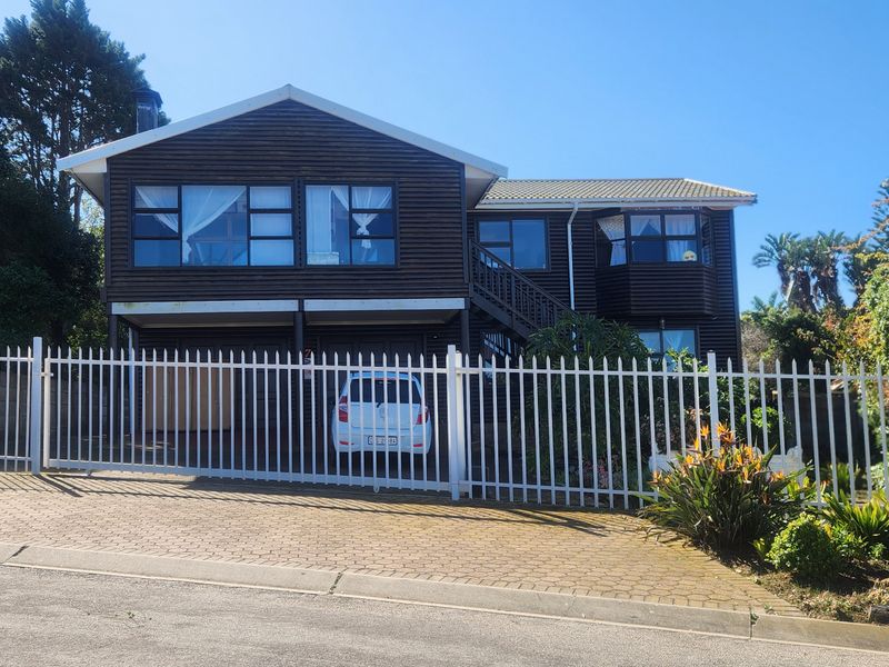 Front elevation of a two-story timber-clad house featuring a paved driveway, metal security fencing, and a gabled roof structure.