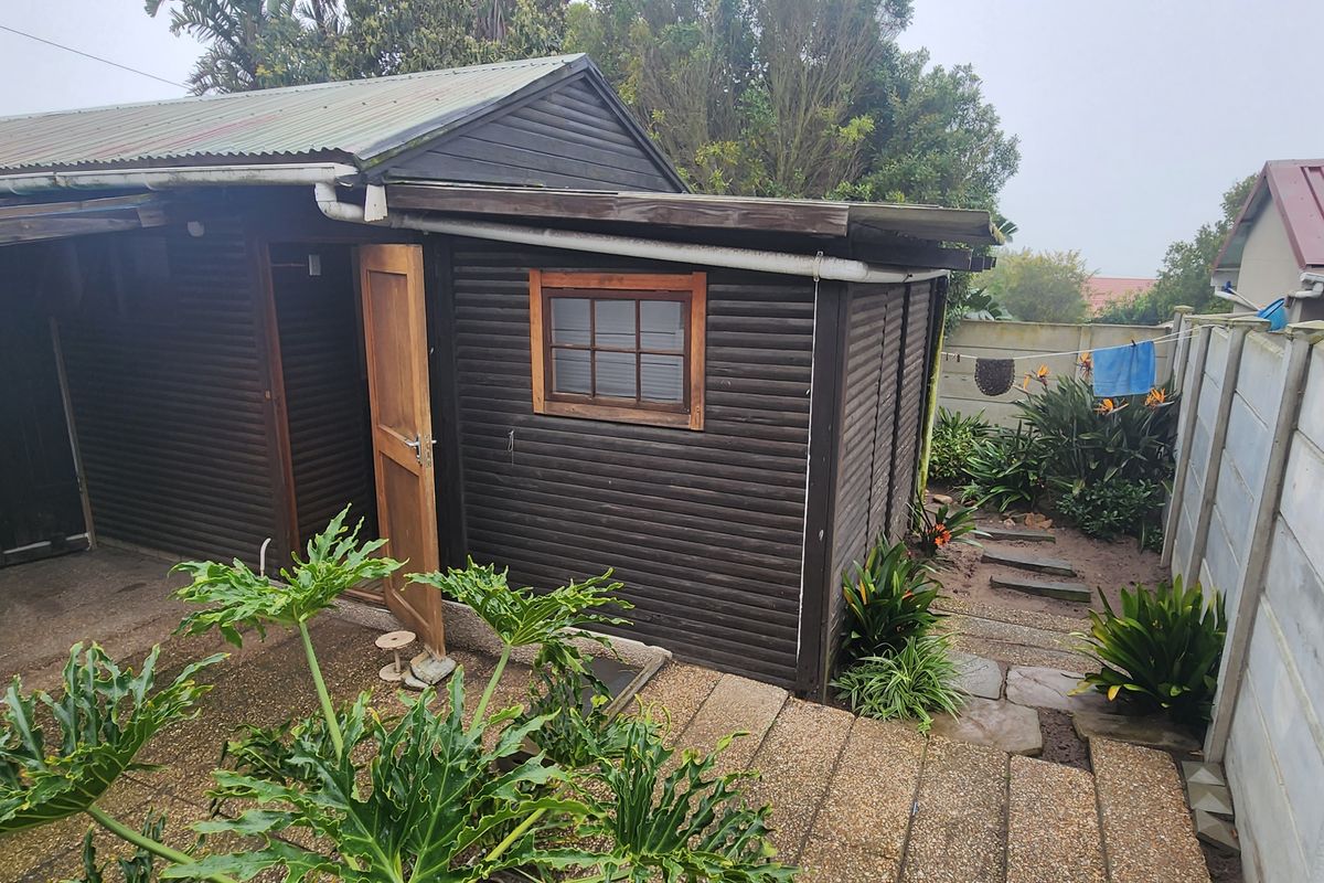 Rear exterior view of a timber-clad structure with a corrugated metal roof, featuring a paved patio area and precast concrete boundary wall.