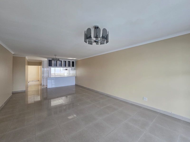 An open-plan living and kitchen area featuring neutral tiled flooring, modern cabinetry, and a breakfast counter, presented in a clean, vacant state.