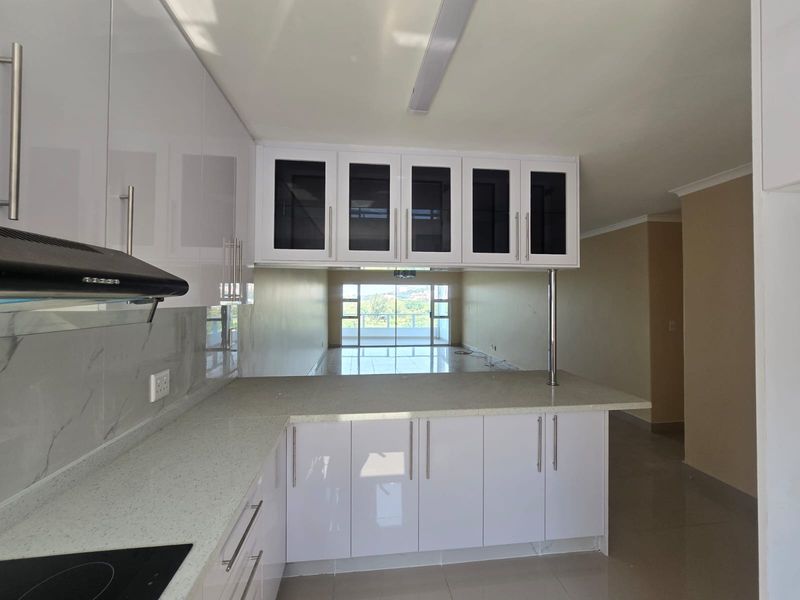 Modern kitchen featuring glossy white cabinetry, stone countertops, and a breakfast bar, overlooking an open-plan living area with balcony access.
