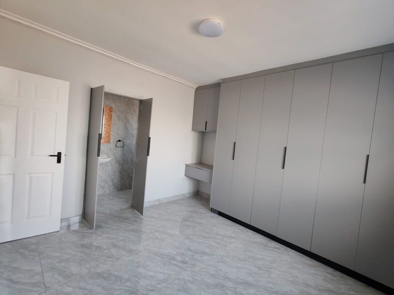 A bright, modern bedroom featuring neutral tiled flooring, extensive built-in grey cabinetry, and direct access to an en-suite bathroom.