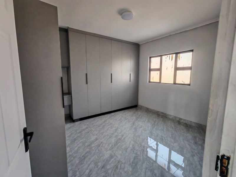 A clean, empty bedroom featuring modern grey built-in cupboards, neutral tiled flooring, and a window allowing natural light.
