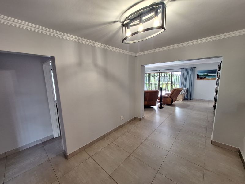 An open-plan living area featuring neutral tiled flooring, decorative ceiling cornices, and a contemporary pendant light fixture, leading to a bright rear living space.