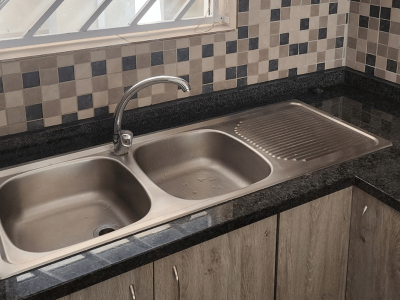 A clean kitchen area featuring a stainless steel double sink, granite countertops, and a modern mosaic tile backsplash with light wood-finish cabinetry.