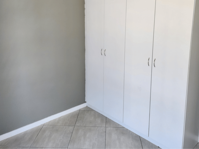 A clean, empty bedroom corner featuring neutral grey walls, white built-in cupboards, and light-colored tiled flooring.