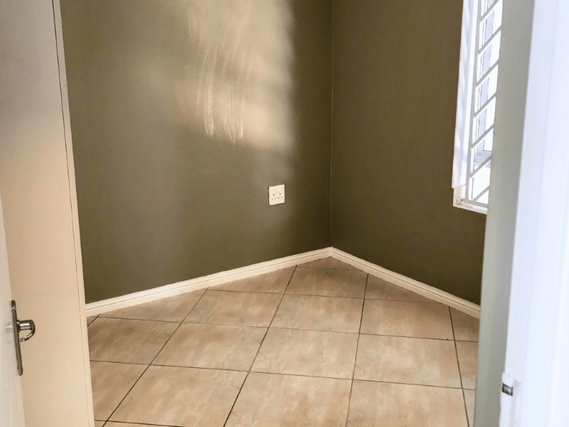 An empty bedroom featuring neutral tiled flooring, olive-toned walls, and a window fitted with security bars.