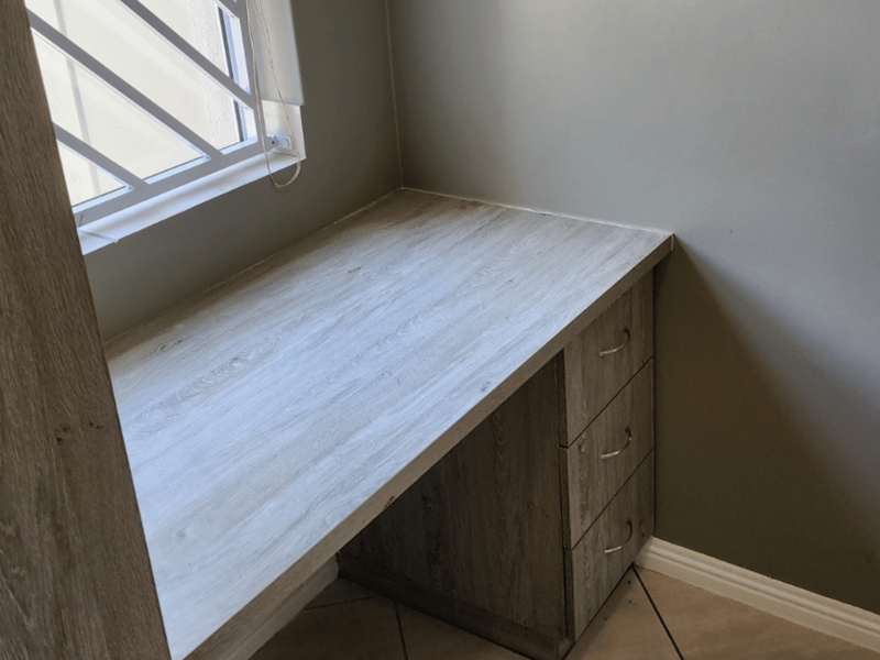 A compact home office space featuring a built-in desk with integrated drawer storage and security bars on the window.