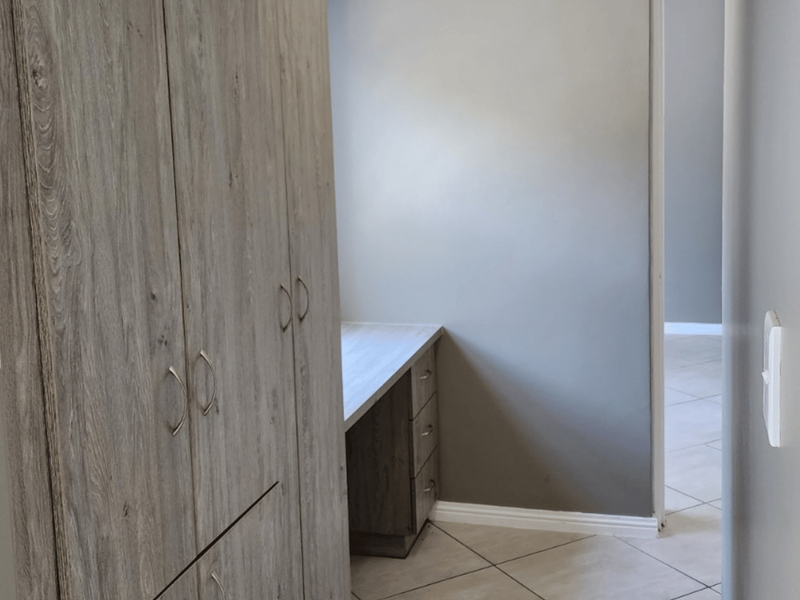 A clean bedroom featuring modern wood-look built-in cupboards and a matching integrated desk, finished with neutral tiled flooring.