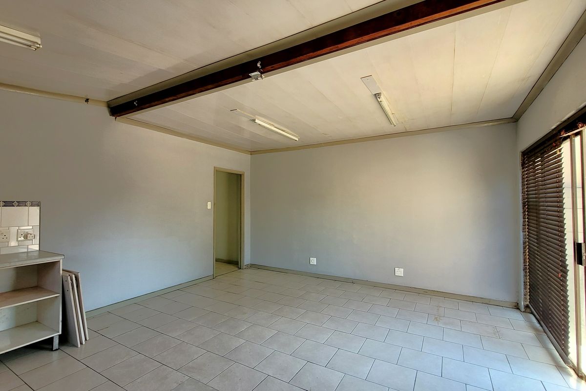 An empty open-plan living area featuring tiled flooring, an exposed timber ceiling beam, and a sliding glass door with venetian blinds.