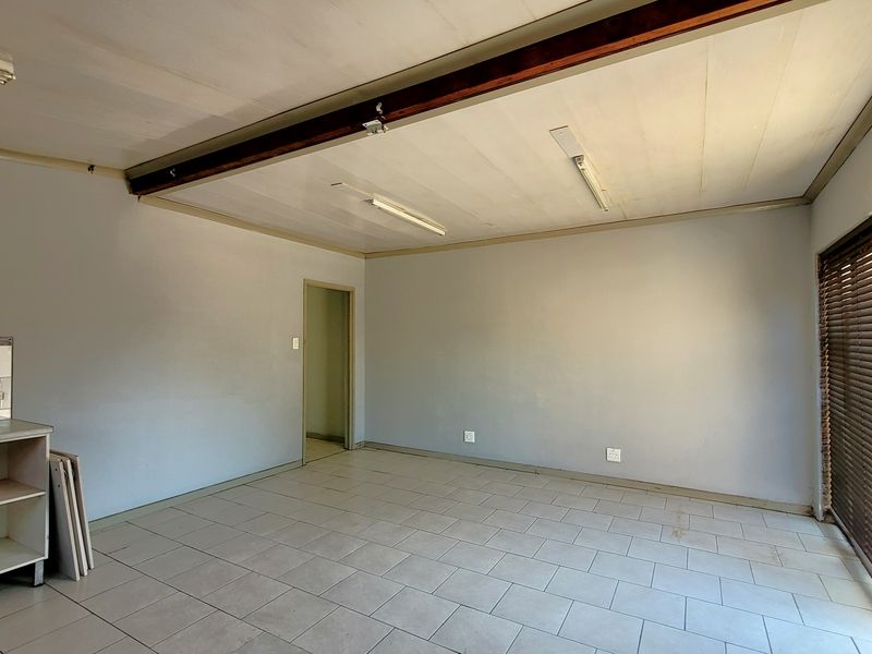 An empty open-plan living area featuring tiled flooring, an exposed timber ceiling beam, and a sliding glass door with venetian blinds.