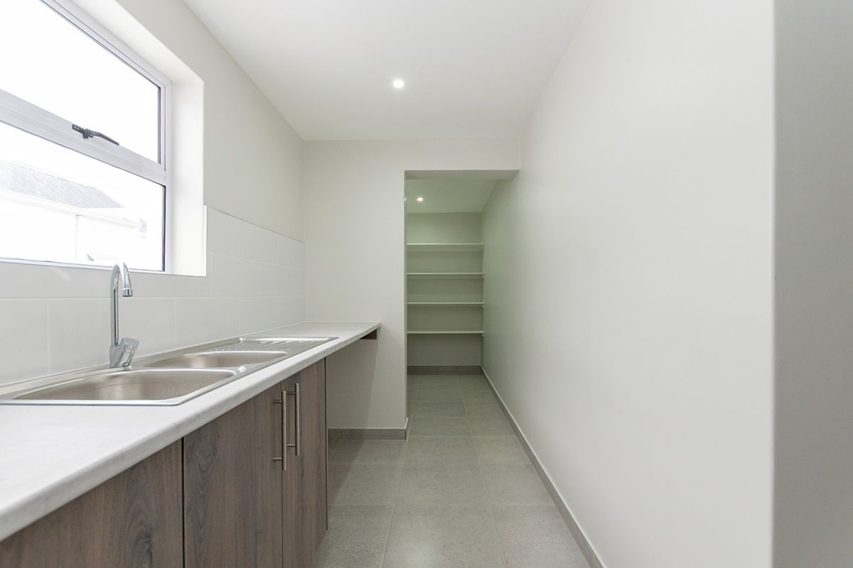 A clean, modern scullery featuring a double sink, laminate countertops, ample built-in cabinetry, and a separate walk-in pantry area with fixed shelving.