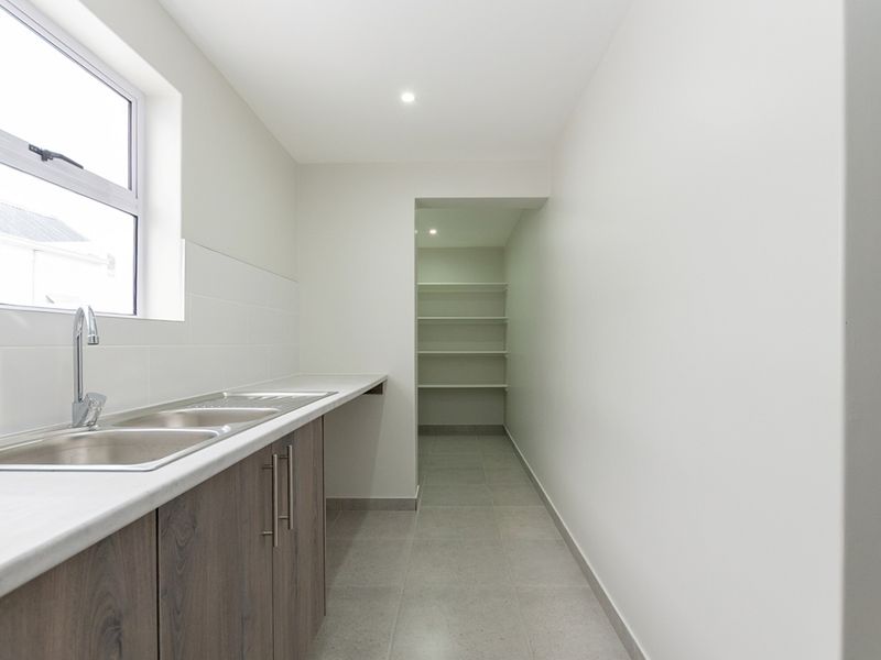 A clean, modern scullery featuring a double sink, laminate countertops, ample built-in cabinetry, and a separate walk-in pantry area with fixed shelving.