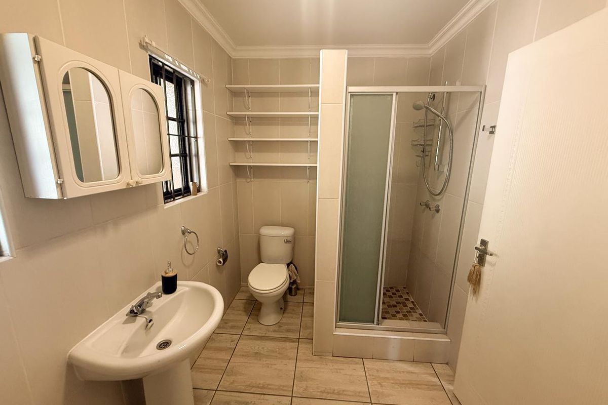 A functional bathroom featuring a pedestal basin, a separate shower enclosure, a toilet, and wall-mounted shelving with neutral tiled finishes throughout.