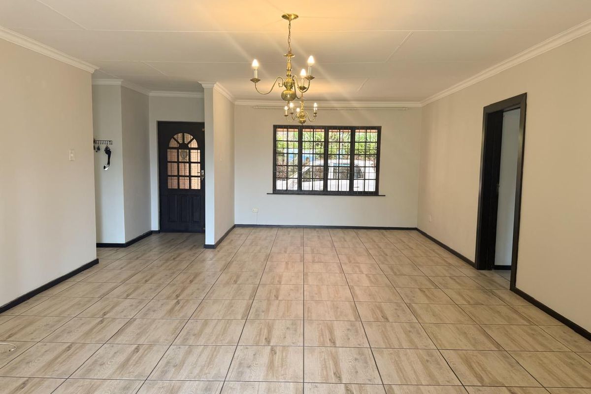 A spacious, empty living area featuring wood-look tiled flooring, coved cornicing, and a classic brass chandelier, providing a clean and neutral blank canvas.