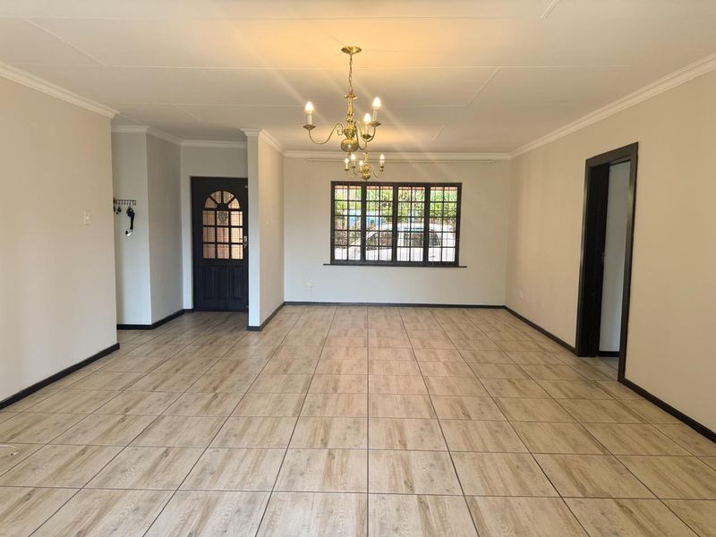 A spacious, empty living area featuring wood-look tiled flooring, coved cornicing, and a classic brass chandelier, providing a clean and neutral blank canvas.