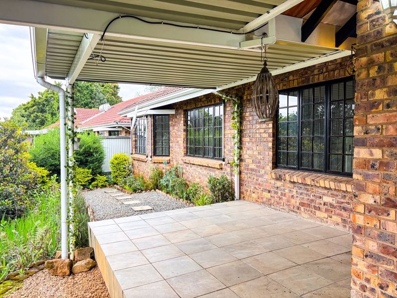 A covered outdoor patio featuring tiled flooring, exposed brick walls, and black metal-framed windows, overlooking a landscaped garden area.