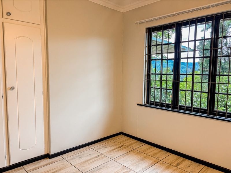 A bright, empty bedroom featuring tiled flooring, a built-in wardrobe, and a window fitted with security bars, offering a pleasant outdoor view.