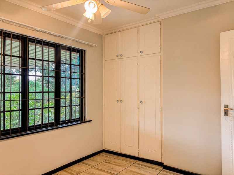 A bright, empty bedroom featuring built-in cupboards, tiled flooring, a ceiling fan, and security bars on the window.