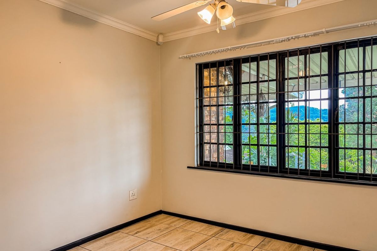 A bright, empty bedroom featuring tiled flooring, a ceiling fan, and large windows fitted with security bars, offering a pleasant outdoor view.