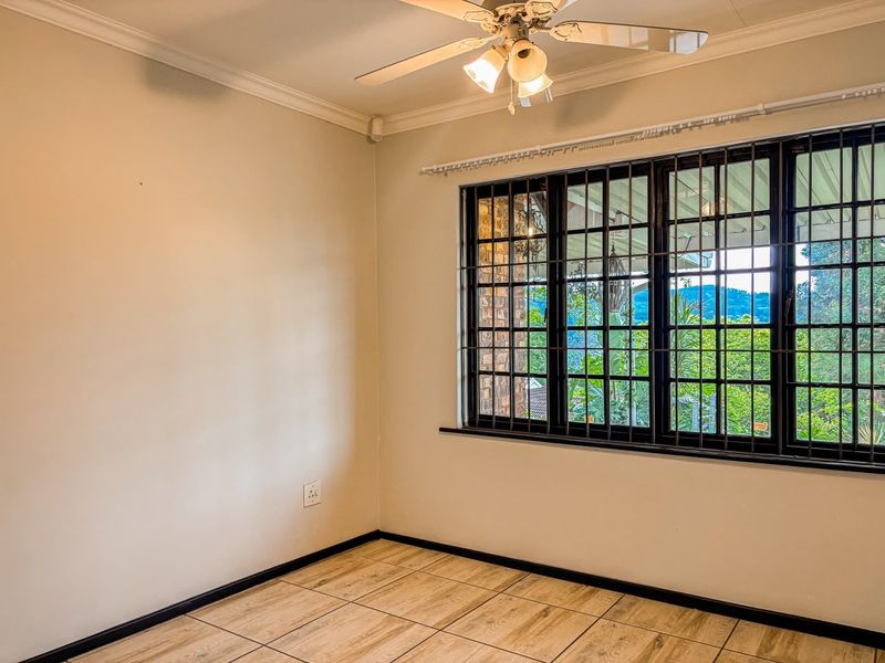 A bright, empty bedroom featuring tiled flooring, a ceiling fan, and large windows fitted with security bars, offering a pleasant outdoor view.