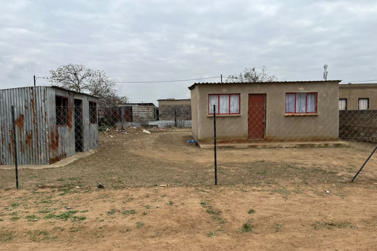 A view of a property featuring a small concrete block building and a separate corrugated metal structure enclosed by a chain-link fence.