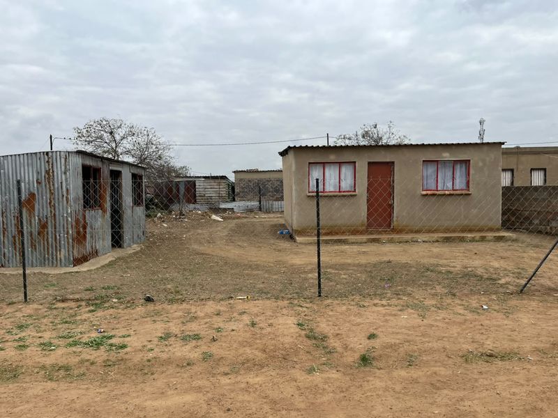 A view of a property featuring a small concrete block building and a separate corrugated metal structure enclosed by a chain-link fence.